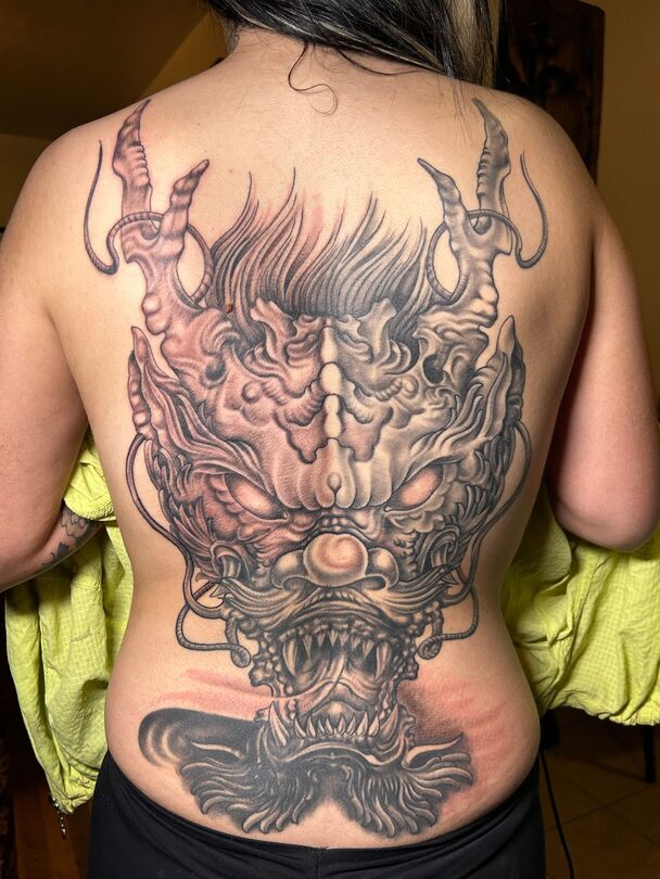 Tattoo of a detailed, gray and red oni mask covers a person's back, with a smaller design on their lower back.