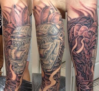 Arm tattoo of a detailed Mesoamerican mask surrounded by stylized leaves and patterns in black and gray.