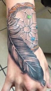 Dreamcatcher and feather tattoo on wrist, detailed gray and brown tones, with colorful beads.