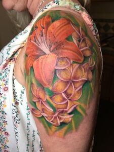 Tattoo of orange lily and yellow plumeria flowers on a person's upper arm, with green leaves and a floral shirt.