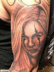 Tattoo of a person with blonde hair and clown-like makeup on their arm. The art is in black and gray.