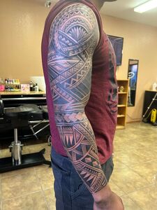 Man with full arm tribal tattoo, wearing maroon shirt and dark jeans, standing in tattoo parlor.