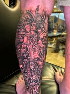 Tattoo of a person in a headdress, surrounded by architectural structures and sunburst, on a leg.