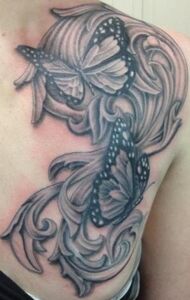 Black and white tattoo of butterflies and ornate scrolling design on shoulder.
