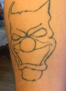 Outline of a clown face tattoo on skin, with horns and an angry expression.
