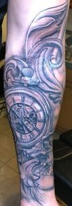 Tattoo of a pocket watch with Roman numerals, surrounded by ornate swirls, on a person's arm.