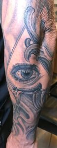 Tattoo of a detailed eye within a triangle and swirling gray designs on an arm.