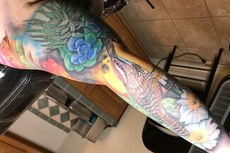 Colorful arm tattoo featuring flora, a bird, and blue sky.
